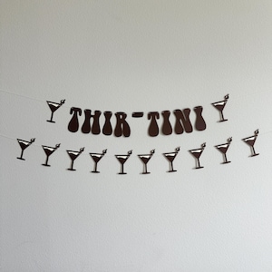 May include: Black and white paper banner with the text "Thir-tini" and a series of martini glasses. The banner is strung together with white string.