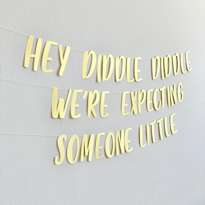 May include: Gold banner with the phrases "HEY DIDDLE DIDDLE", "WE'RE EXPECTING", and "SOMEONE LITTLE" strung across a white wall. The banner is made of gold cardstock and is a decorative item.