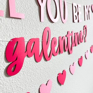 May include: A Valentine's Day banner with the words "Will You Be My Galentine?" in pink script. The banner includes pink and red heart-shaped cutouts strung along a white string, creating a festive decoration.