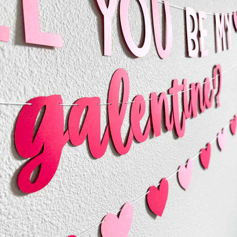 May include: A Valentine's Day banner with the words "Will You Be My Galentine?" in pink script. The banner includes pink and red heart-shaped cutouts strung along a white string, creating a festive decoration.