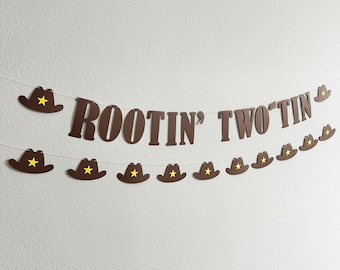 May include: A brown banner with the text "Rootin' Two Tin" and cowboy hat cutouts with yellow stars. The banner is strung on a white string and is against a white wall.