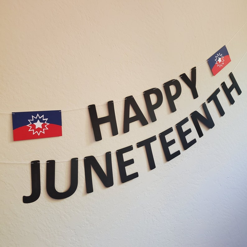 May include: Black paper banner with the words "Happy Juneteenth" in white letters. Two small blue and red flags with a white star are hanging from the banner.