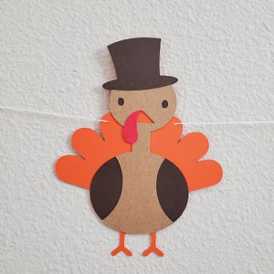 May include: A paper craft turkey with a brown hat, orange feathers, and a red beak. The turkey is hanging from a white string.