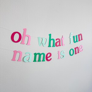 Puede incluir: Una pancarta de papel colorida con la frase "Oh what fun name is one" en letras rosas, verdes y blancas. La pancarta es perfecta para una fiesta de primer cumplea&ntilde;os.