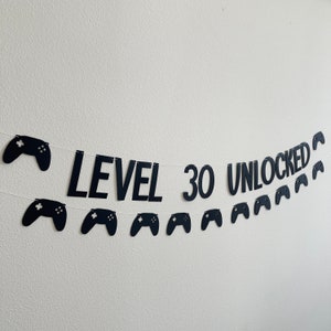 May include: Black banner with the text "LEVEL 30 UNLOCKED" and controller shapes. The banner is strung on a white string and is against a white wall. The banner is a birthday decoration.