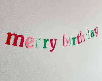 May include: A festive banner with the words "merry birthday" in pink, red, and green letters. The letters are cut out of paper and strung together with white thread.