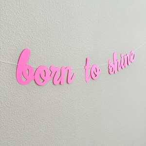 May include: A pink banner with the words "born to shine" in a cursive font. The banner is strung on a white string and hangs against a light gray wall. The letters are cut out and have a smooth finish.