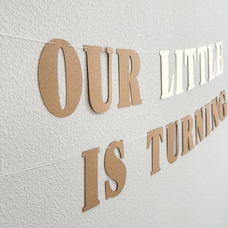 May include: A banner with the words "OUR LITTLE IS TURNING" in brown and white letters strung on a white string. The banner is hanging against a textured, light gray wall.