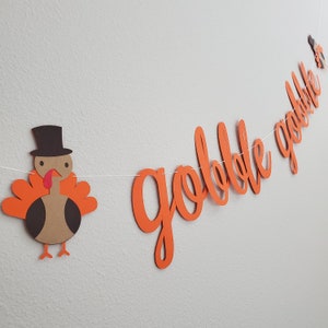 May include: Orange paper banner with the word "gobble gobble" in a cursive font. A brown paper turkey with a black top hat is hanging from the banner.