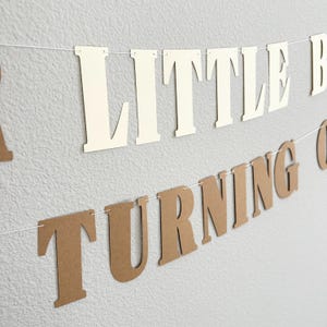 May include: A banner with the words "LITTLE B" in cream-colored letters and "TURNING ONE" in brown letters. The letters are strung on a white string and hang against a textured gray wall.
