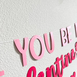 May include: A pink banner with the words "YOU BE MINE" and "Valentine's" in a cursive font. The banner is strung on a white string and is against a white textured wall.