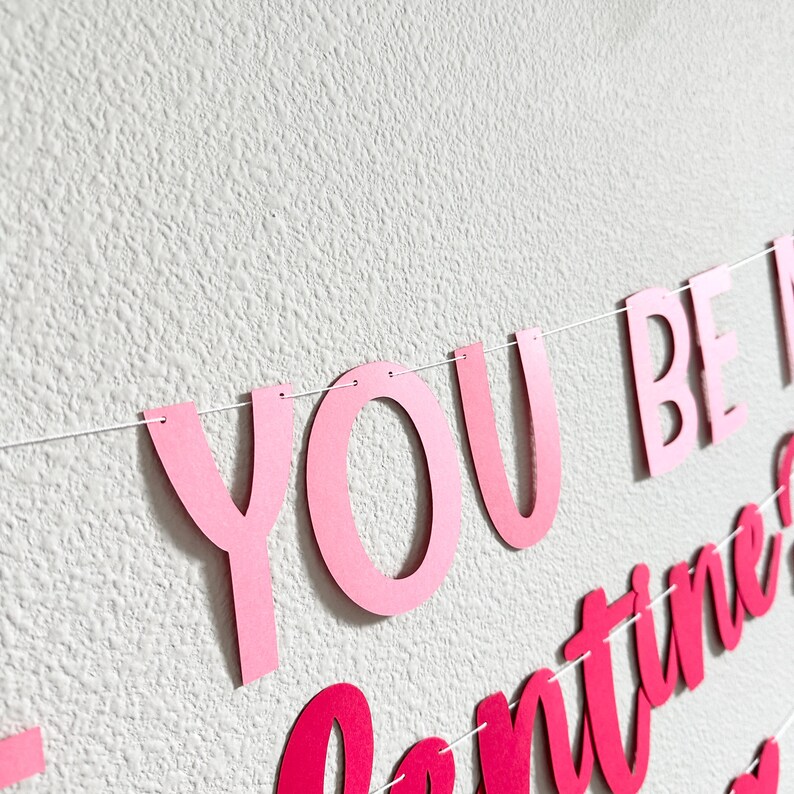 May include: A pink banner with the words "YOU BE MINE" and "Valentine's" in a cursive font. The banner is strung on a white string and is against a white textured wall.
