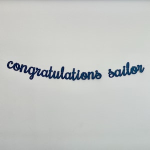May include: A navy blue banner with the words "congratulations sailor" in a cursive font. The banner is strung on a thin white string and is set against a white background.