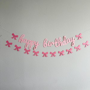 May include: Pink paper banner with the words "happy birthday" and pink paper bows strung on a white string.