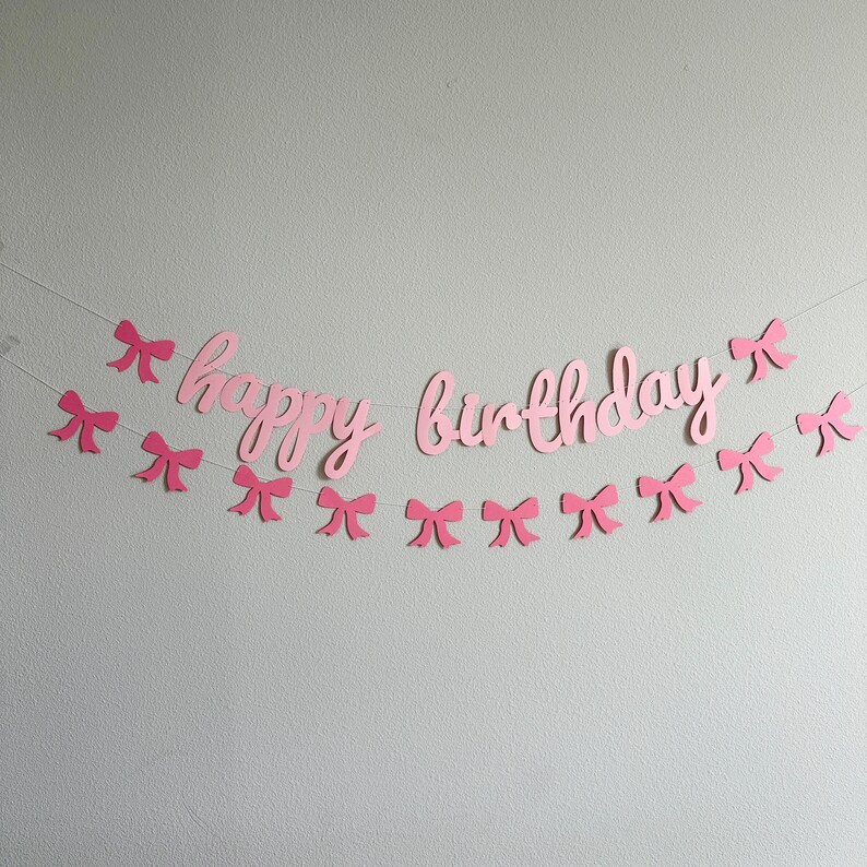 May include: Pink paper banner with the words "happy birthday" and pink paper bows strung on a white string.
