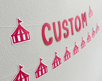 May include: A custom banner with the word "CUSTOM" in red, strung with small circus tent cutouts. The banner is against a light gray wall, creating a festive and personalized decoration.