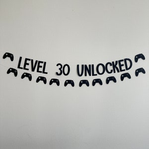 May include: Black banner with the text "LEVEL 30 UNLOCKED" and black controller cutouts. The banner is strung on a white string and is against a white wall. A birthday party decoration.