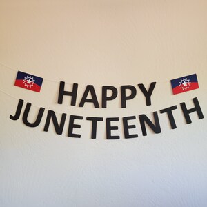 May include: A black banner with the words "Happy Juneteenth" hanging on a white wall. Two small flags with a red, white, and blue star design are hanging on either side of the banner.
