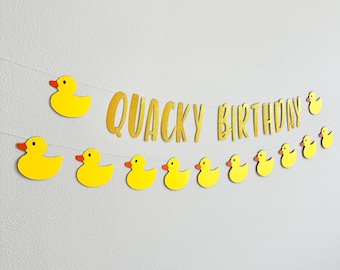 May include: A gold glitter banner with the words "Quacky Birthday" and yellow rubber duck decorations.