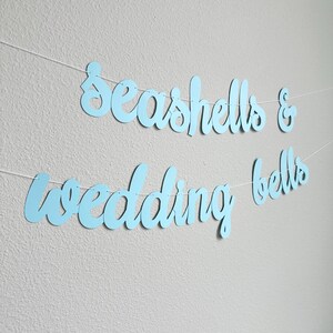 May include: A light blue banner with the words "Seashells & Wedding Bells" in a cursive font. The banner is strung on a thin white string and hangs against a light gray wall.