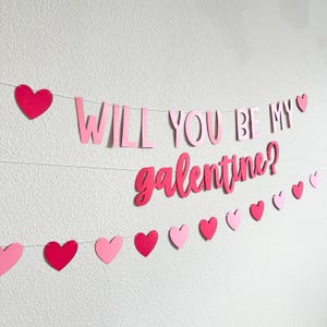 May include: A pink and red paper banner with the words "WILL YOU BE MY galentine?" strung across a white wall. The banner includes heart-shaped cutouts in varying shades of pink and red.