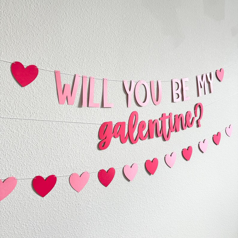 May include: A pink and red paper banner with the words "WILL YOU BE MY galentine?" strung across a white wall. The banner includes heart-shaped cutouts in varying shades of pink and red.