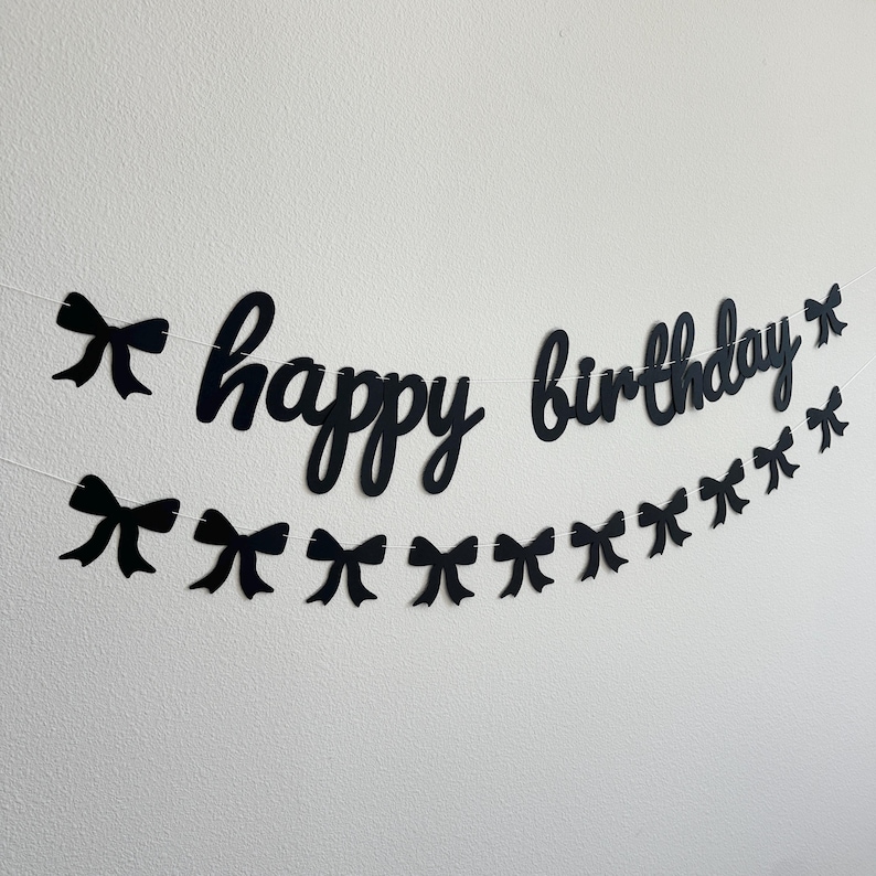 Pode incluir: Faixa de feliz anivers&aacute;rio em preto e branco com decora&ccedil;&otilde;es de la&ccedil;o preto. A faixa est&aacute; pendurada em uma parede branca.