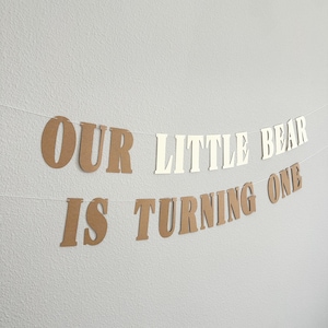 May include: A banner with the text "OUR LITTLE BEAR IS TURNING ONE" in brown and white letters. The banner is strung on a white string and is hanging against a neutral wall.