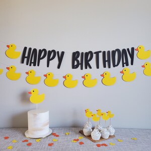 May include: A yellow and black birthday banner with the words "Happy Birthday" and yellow rubber duck decorations. A white cake with a yellow rubber duck on top is on a table with yellow and orange confetti.