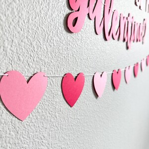 May include: A Valentine's Day banner with the words "Will you be my Valentine?" in pink script. A string of pink paper hearts hangs below. The hearts vary in shade from light to dark pink, creating a festive decoration.