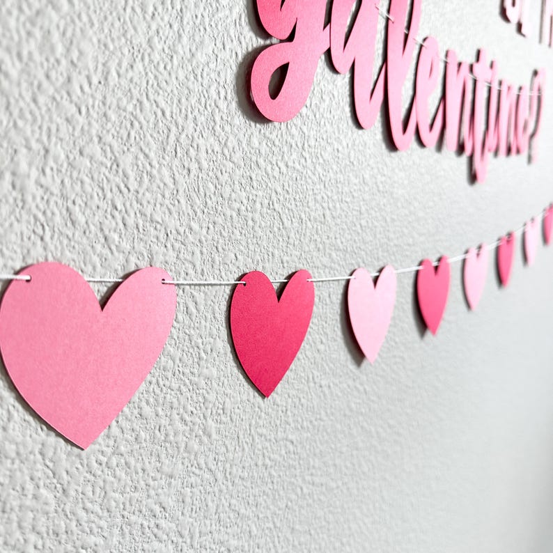 May include: A Valentine's Day banner with the words "Will you be my Valentine?" in pink script. A string of pink paper hearts hangs below. The hearts vary in shade from light to dark pink, creating a festive decoration.