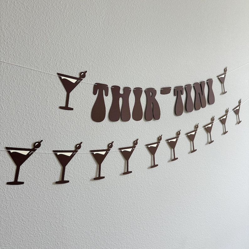 May include: A brown paper banner with the words "Thir-Tini" in a retro font. The banner is decorated with white martini glass cutouts.
