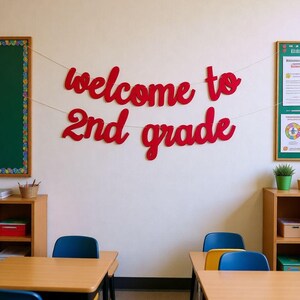 May include: A red script-style wall banner made of individual cut-out letters spelling out the phrase "welcome to 2nd grade" suspended on two thin white strings against a plain off-white wall in a classroom setting.