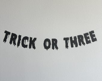 May include: Black and white banner with the text "Trick or Three" in dripping letters. The banner is hanging on a white wall.