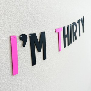 May include: A pink and black banner with the text "I'M THIRTY". The letters are cut out of paper and strung together with white string.