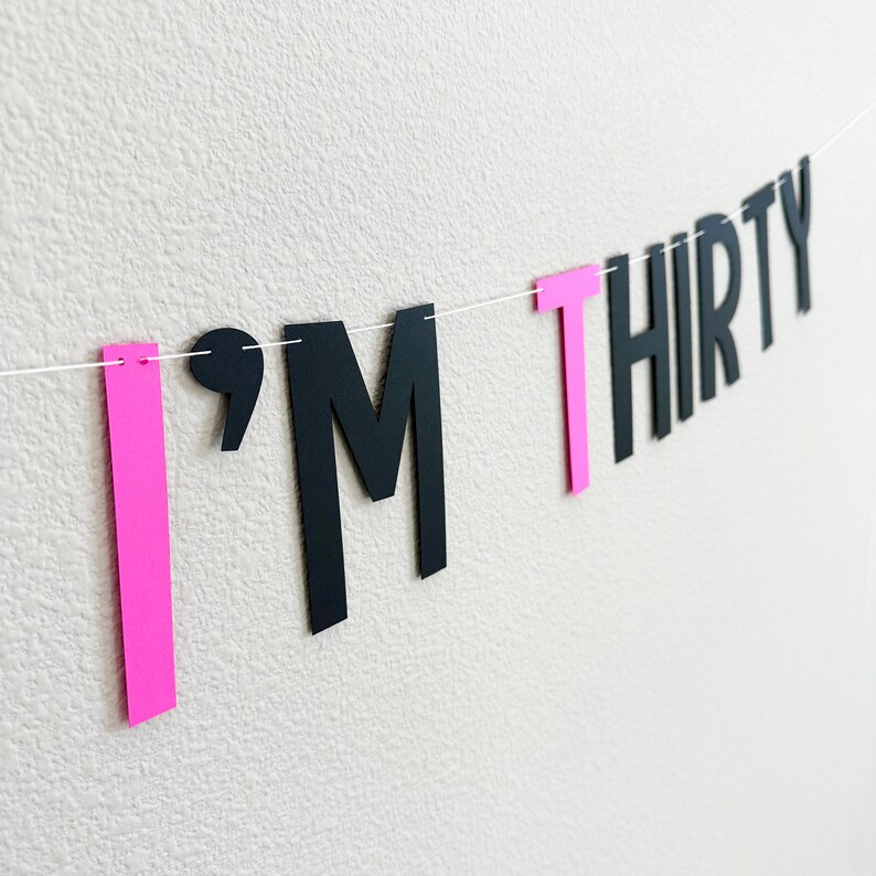 May include: A pink and black banner with the text "I'M THIRTY". The letters are cut out of paper and strung together with white string.