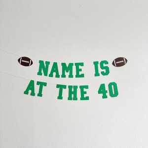 Puede incluir: Pancarta verde y blanca con el texto "NAME IS AT THE 40" y dos balones de f&uacute;tbol marrones en cada extremo.