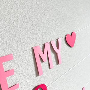 May include: Pink paper banner with the words "BE MY" and a heart shape strung on white string. The banner is against a textured white wall.