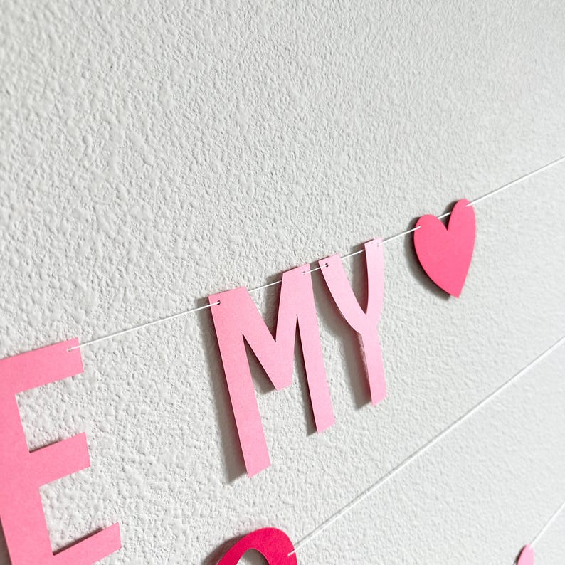 May include: Pink paper banner with the words "BE MY" and a heart shape strung on white string. The banner is against a textured white wall.