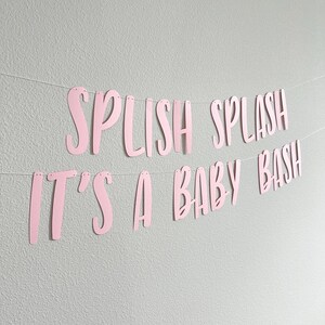 May include: Pink paper banner with the words "Splish Splash" and "It's a Baby Bash" strung on a thin white string. The banner is hanging against a light gray wall.