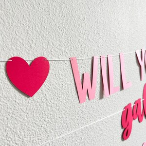 May include: A pink heart-shaped banner with the words "WILL YOU" in pink letters. The banner is strung on a white string and is against a white textured wall. The banner is a decorative item.