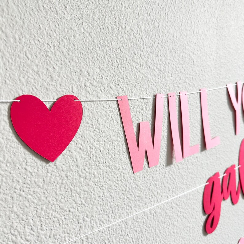 May include: A pink heart-shaped banner with the words "WILL YOU" in pink letters. The banner is strung on a white string and is against a white textured wall. The banner is a decorative item.