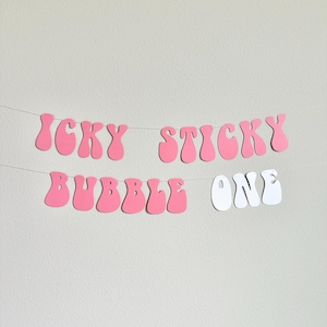 May include: Pink and white banner with the words "ICKY STICKY BUBBLE ONE" strung across a thin wire. The letters are in a retro, bubble-style font, with the words "ICKY STICKY BUBBLE" in pink and "ONE" in white. The banner is against a plain, light gray background.