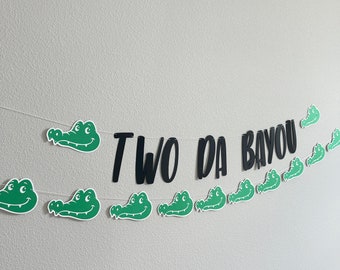 May include: A green and black alligator themed banner with the text "TWO DA BAYOU". The banner is made of paper and has a string running through it.