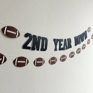 May include: A black and white banner with the text "2nd Year Down" and football-shaped cutouts. The banner is strung with white string and is hanging on a white wall.