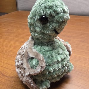 May include: A plush, green turtle with a textured, fuzzy exterior. The turtle has a light brown shell and large, black eyes. The stuffed animal is sitting on a wooden surface.