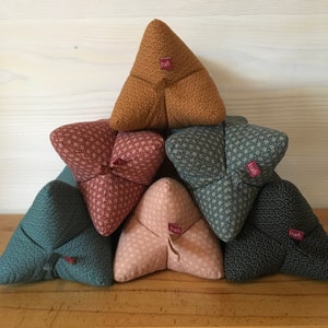 Könnte beinhalten: Eine Pyramide aus dreieckigen Kissen in verschiedenen Farben, darunter Gold, Türkis und Rosa. Jedes Kissen hat einen gemusterten Stoff mit sich wiederholendem Design und einem kleinen Etikett mit der Aufschrift "bafi".