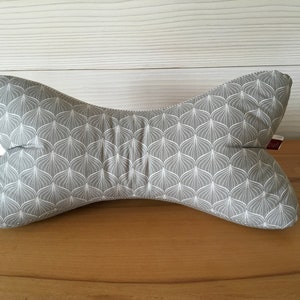 Könnte beinhalten: Ein Nackenkissen mit grauem und weißem Muster. Das Kissen hat eine einzigartige Form und ein sich wiederholendes blattartiges Design. Es besteht aus weichem Stoff und soll Nacken und Schultern stützen. Es ist etwa 40 cm breit.