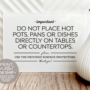 Op de afbeelding: Een wit bord met zwarte tekst die luidt: "Belangrijk. Plaats geen hete potten, pannen of schotels direct op tafels of aanrechtbladen. Gebruik de meegeleverde onderzetters. Dank u!" Het bord staat op een houten oppervlak met een witte vaas en gedroogde bloemen.