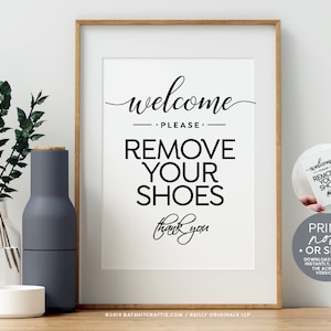 May include: A black and white printable sign that says "Welcome Please Remove Your Shoes Thank You". The sign is framed in a light brown wood frame and is sitting on a wooden table with a vase of greenery and a gray bottle.